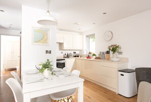 A modern kitchen with white cabinetry, a dining area with a white table set for two, and a large window providing natural light. - A stunning ground floor apartment with a large outdoor terrace and sea views. (Carbis Bay)