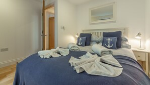Ein ordentlich bezogenes Bett mit dunkelblauer Bettwäsche und Kissen mit Anker-Motiv, eine Nachttischlampe und ein Nachttisch mit Lampe.