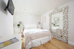 A bedroom with a large ornate mirror, a bed with white bedding, a bedside table with a lamp, and a TV mounted on the wall.