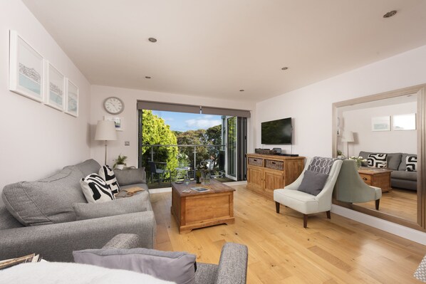 A modern living room with a grey sofa, a wooden coffee table, and a grey armchair. There is a large window with a view of trees and a balcony.