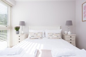 A neatly made bed with white linens, two bedside tables with lamps, and a window with blinds.