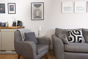 A living room with a grey armchair, a grey sofa, and a side table with a lamp. There are framed artworks on the wall and a wooden shelf with various items.
