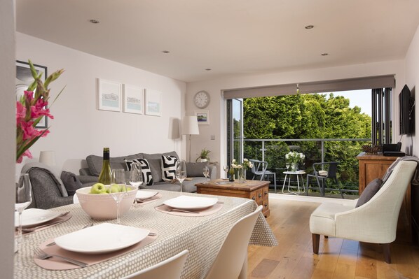 A modern living room with a dining area, a sofa set, a coffee table, and a view of a lush garden through large windows.