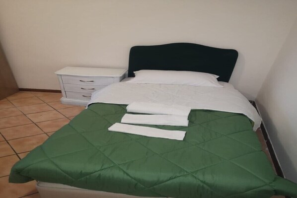 A single bed with a green quilt, white bedding, and a dark headboard in a room with a white nightstand and a drawer unit.