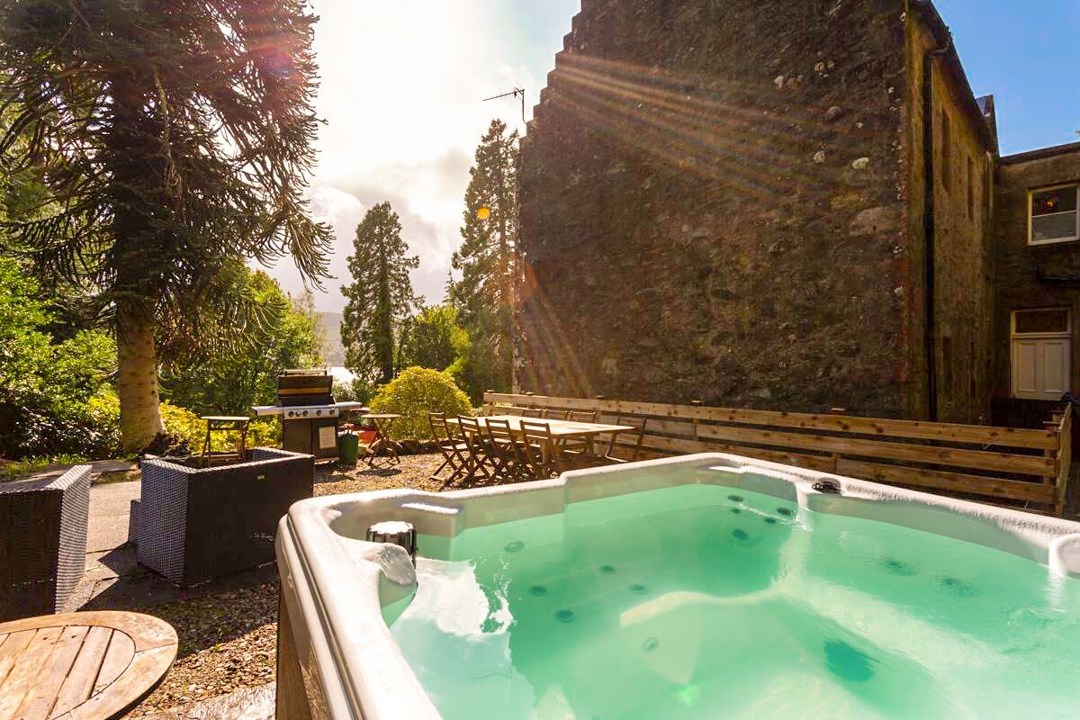A hot tub in a backyard with a stone wall, outdoor furniture, and a grill.