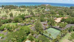 An aerial view of a resort with a tennis court, lush greenery, and a view of the ocean.