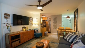 A living room with a sofa, a wooden coffee table, a TV, a ceiling fan, and a kitchen in the background.