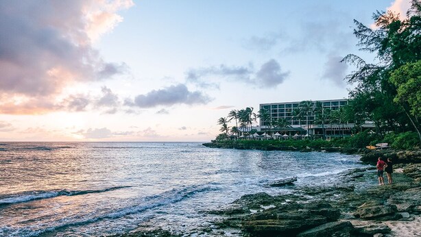 A coastal hotel with a rocky shoreline, palm trees, and a building with multiple balconies overlooking the sea.