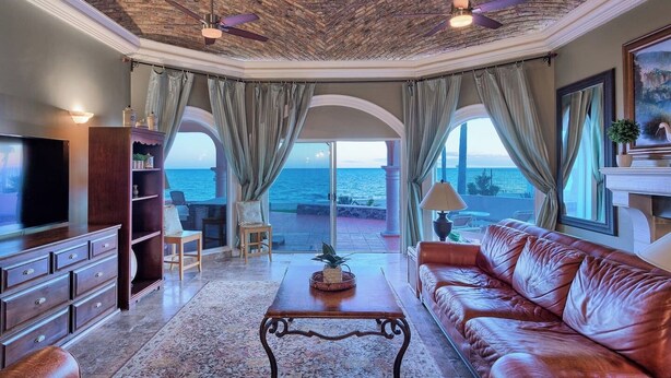 A spacious living room with a leather sofa, a coffee table, and a TV. There are two ceiling fans, a large window with curtains, and a view of the sea.