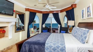 A bedroom with a large bed, a ceiling fan, a TV, and a view of the sea through the windows.