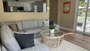 Ein Wohnzimmer mit einem grauen Ecksofa, einem Glastisch und Blick auf den Garten durch eine Schiebetür.