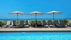 A poolside area with lounge chairs and umbrellas, a clear blue sky, and a view of the ocean.