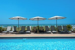 A poolside area with lounge chairs and umbrellas, a clear blue sky, and a view of the ocean. - Twin Palms 1002 by Best Beach Getaways (Panama City Beach)