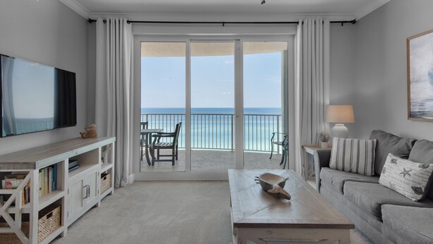 A spacious living room with a large TV, a wooden coffee table, a sofa, and a balcony with a view of the ocean.