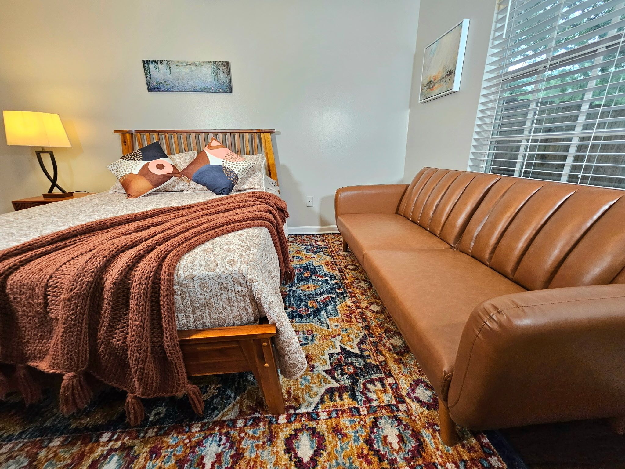 A bedroom with a bed, a brown leather sofa, a patterned rug, and two wall-mounted paintings.
