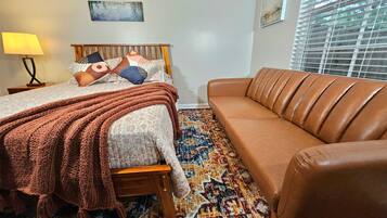 A bedroom with a bed, a brown leather sofa, a patterned rug, and two wall-mounted paintings.
