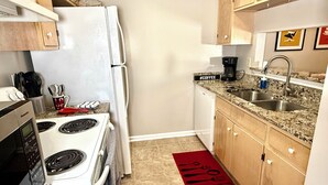 Une cuisine compacte avec un réfrigérateur, une cuisinière et un four à micro-ondes, tous de couleur blanche. On y trouve un comptoir en granit, un évier et des armoires en bois. Un tapis rouge orné d’ustensiles noirs est posé sur le sol.