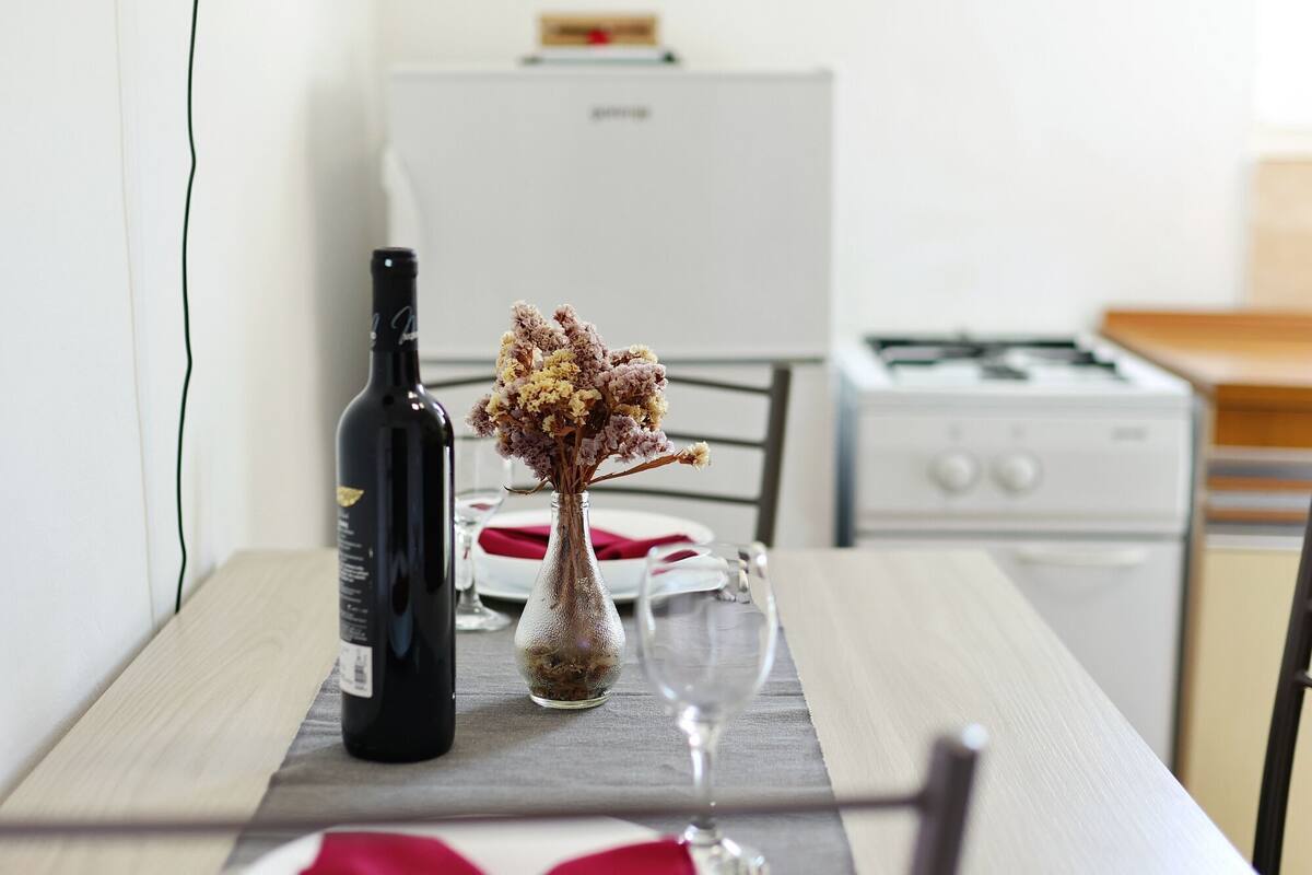 A kitchen with a dining table set for two, a wine bottle, and a vase with dried flowers.