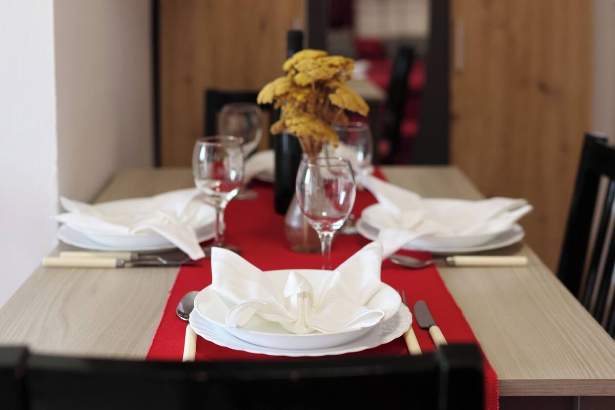 A dining table set with white plates, silverware, and a red table runner.