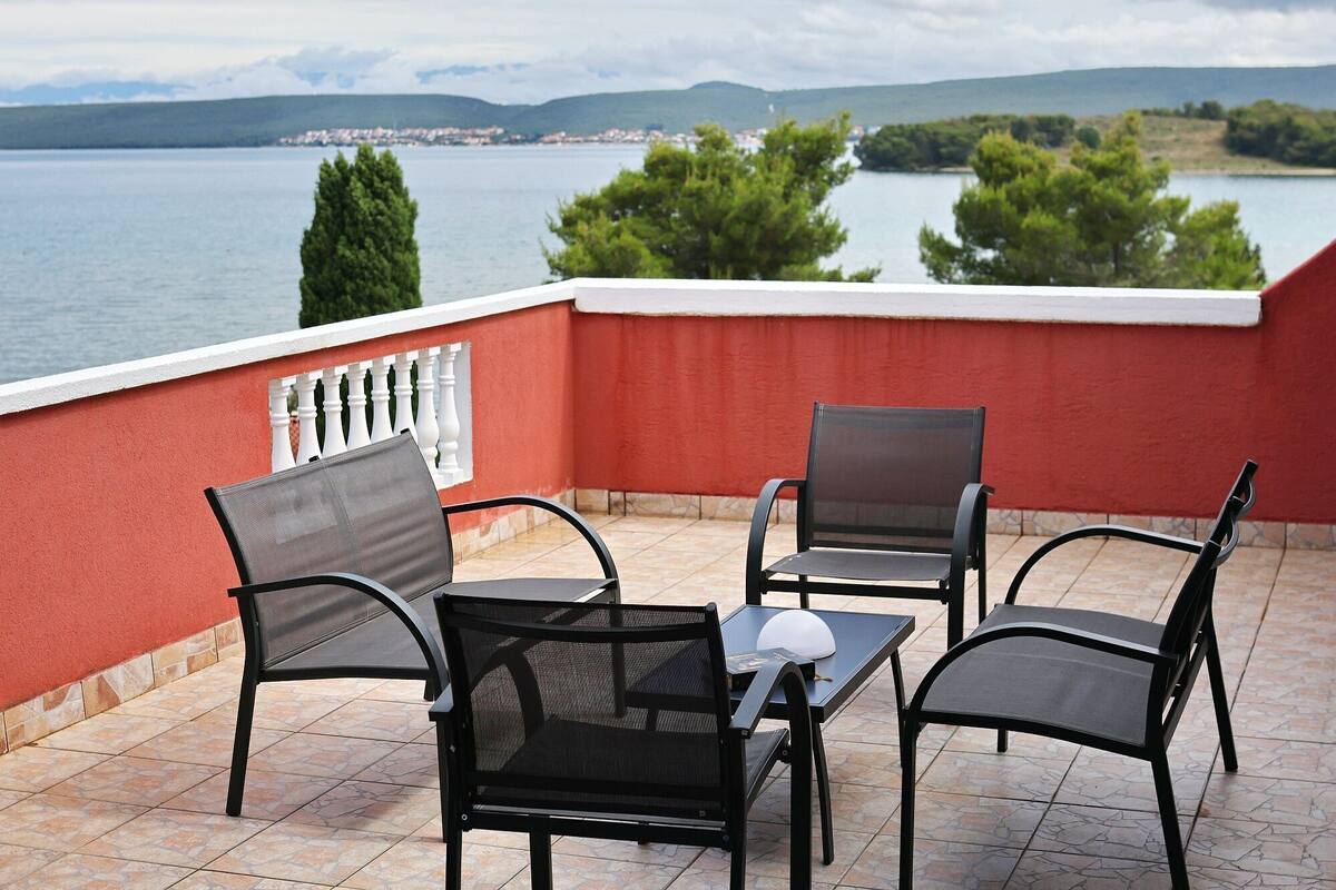 A balcony with two chairs and a table, overlooking a lake with trees and a distant town.