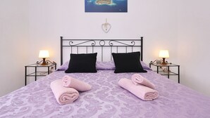 A bed with a purple patterned bedspread, two black pillows, and two pink towels. There are two bedside tables with lamps, a wall clock, and a framed picture of islands. - Apartments Saphron Beach (Pasman)