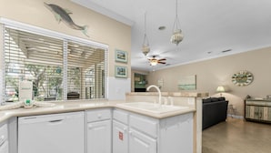 A kitchen with white cabinetry, a built-in dishwasher, a window with blinds, a ceiling fan, and a clock on the wall. - Beach Cottage 120 at Hidden Dunes by Best Beach Getaways (Miramar Beach)
