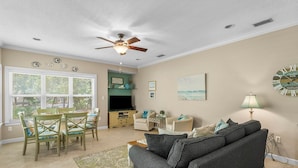 A living room with a gray sofa, a dining table with chairs, a ceiling fan, and a large window with blinds. - Beach Cottage 120 at Hidden Dunes by Best Beach Getaways (Miramar Beach)