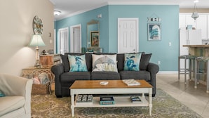 A living room with a sofa, coffee table, and decorative pillows. There is a door, a mirror, and a wall with a picture. - Beach Cottage 120 at Hidden Dunes by Best Beach Getaways (Miramar Beach)