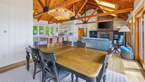Un vaste espace dédié à la salle à manger, avec une grande table et des chaises en bois, une cuisine à l’arrière-plan et un plafond haut avec des poutres apparentes.