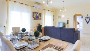 A dining area with a wooden table set for a meal, a sofa in the background, and a television in the adjacent room. - SeaSunrise: Modern, spacious Apt, A/C, WiFi, Near beach (Perama)