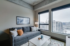 A modern living room with a grey sofa, a framed picture, a lamp, and a view of the city through large windows. - MINNeSTAY* Sable 703 | North Loop | Target Center (Minneapolis)