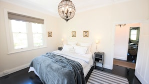 A bedroom with a large bed, a chandelier, a nightstand, and a window with blinds.