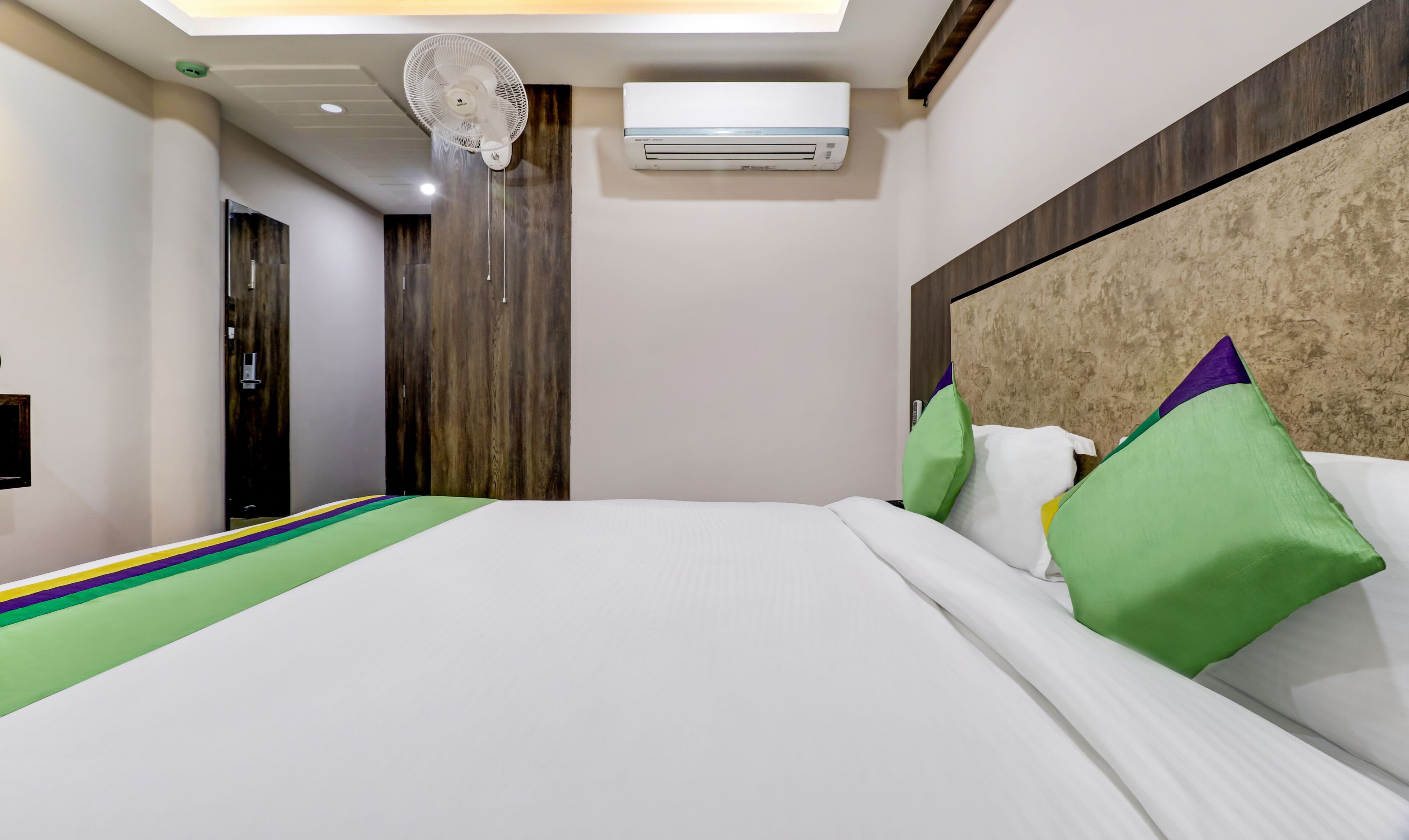 A hotel room with a bed, a ceiling fan, an air conditioning unit, and a wooden wardrobe.