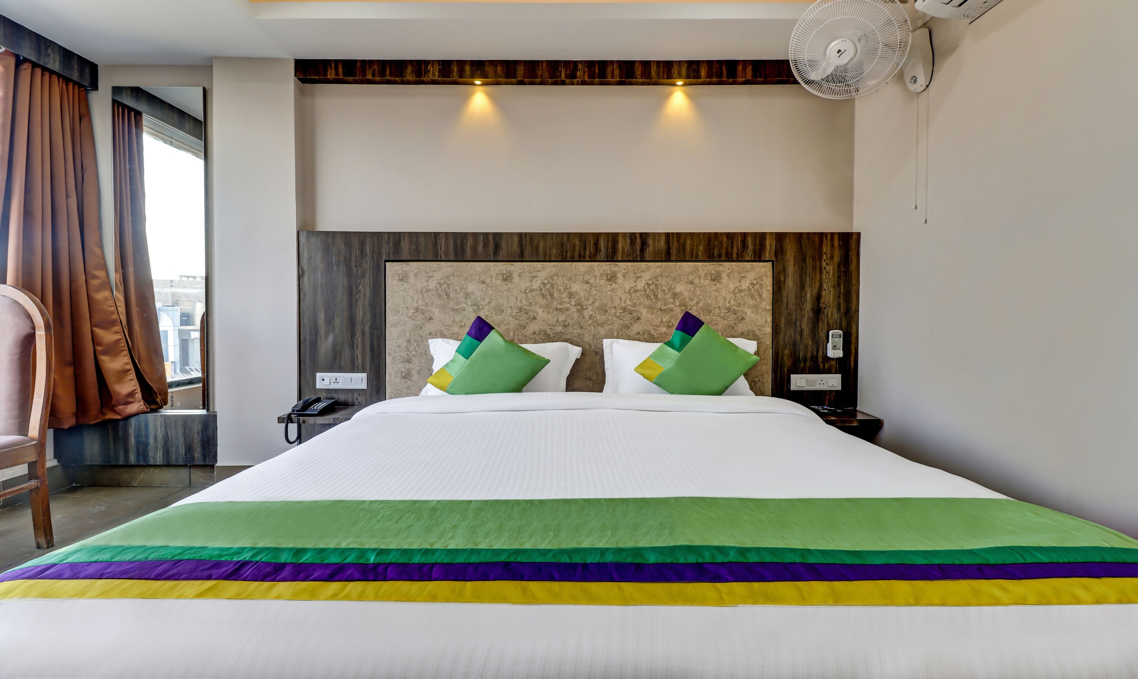 A hotel room with a large bed, two pillows, a bedside table with a phone, a wall-mounted fan, and a window with curtains.