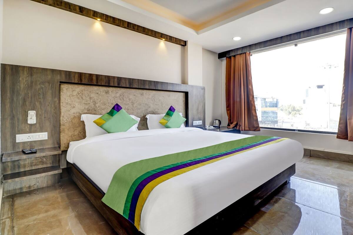 A hotel room with a large bed, a wooden headboard, a nightstand, a window with curtains, and a city view.