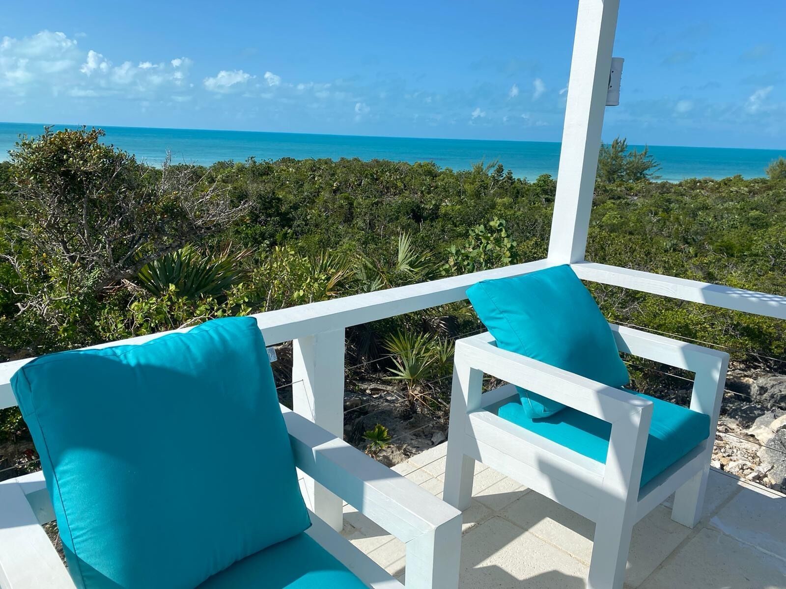 A white outdoor seating area with turquoise cushions, overlooking a tropical landscape and the ocean.