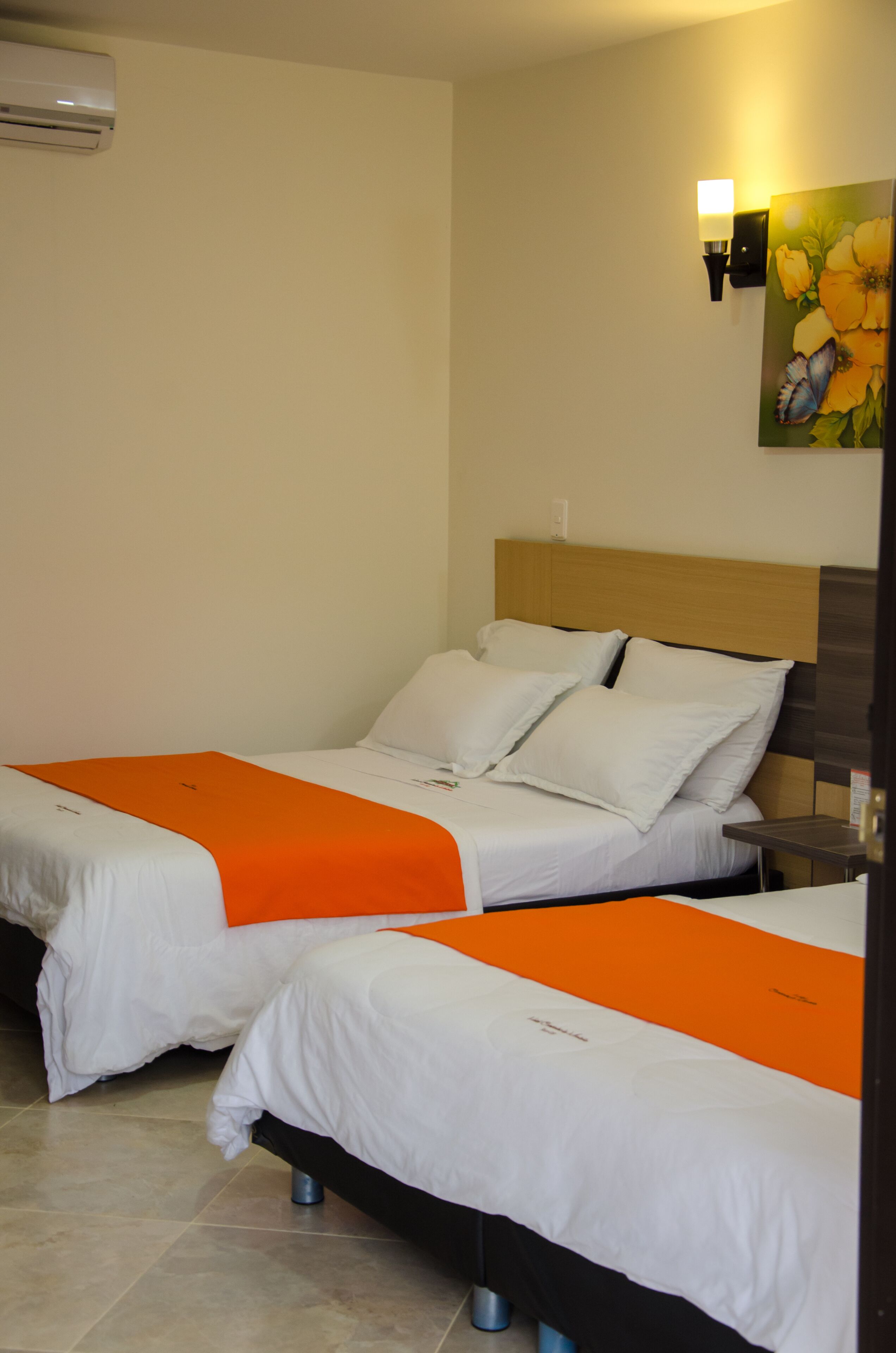 A hotel room with two beds, each with white bedding and orange accents. There is a wall-mounted air conditioner and a painting of flowers and a butterfly on the wall.