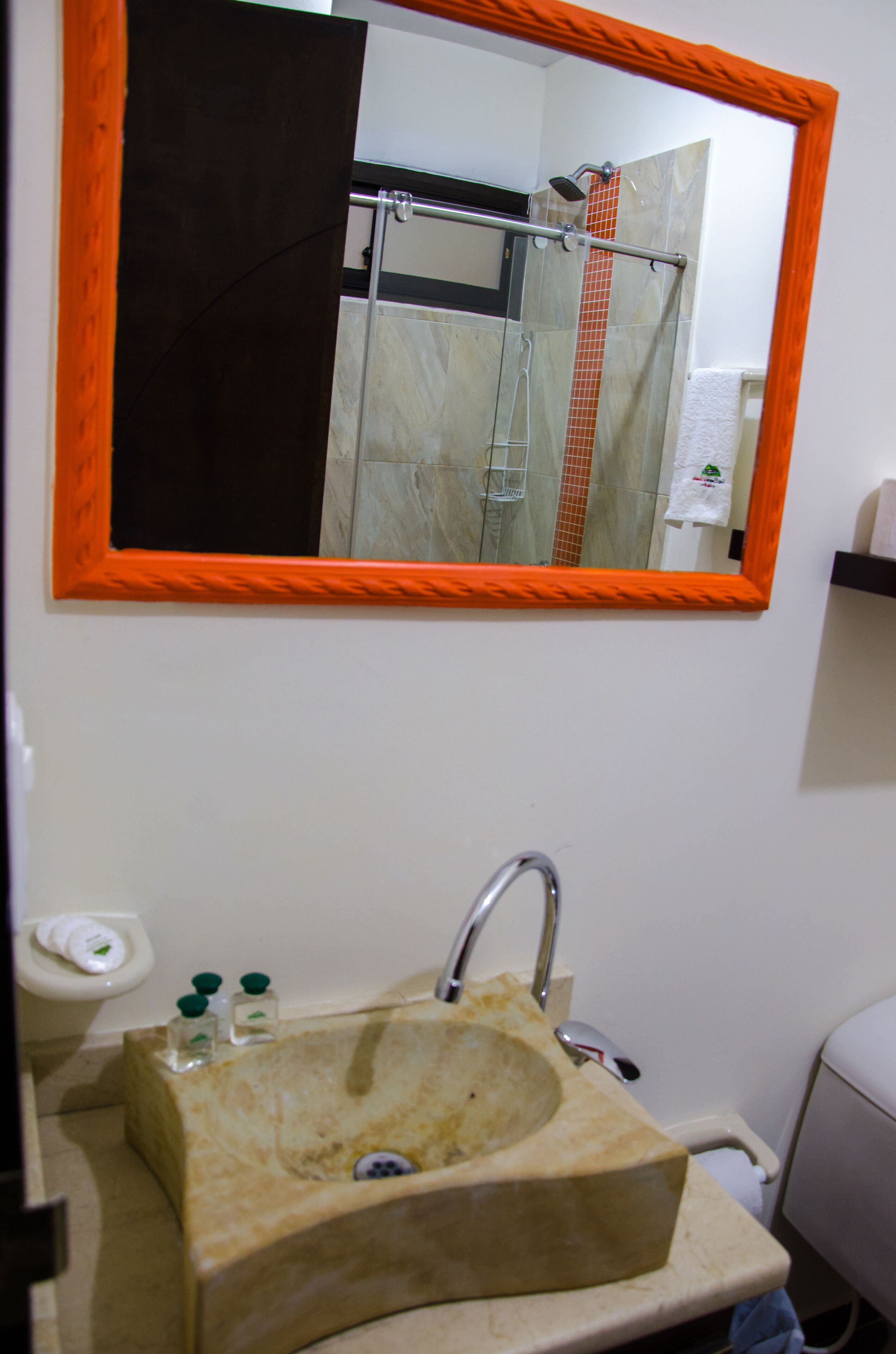 Un baño con un lavamanos color crema, un espejo de marco naranja, y una ducha visible en el reflejo.