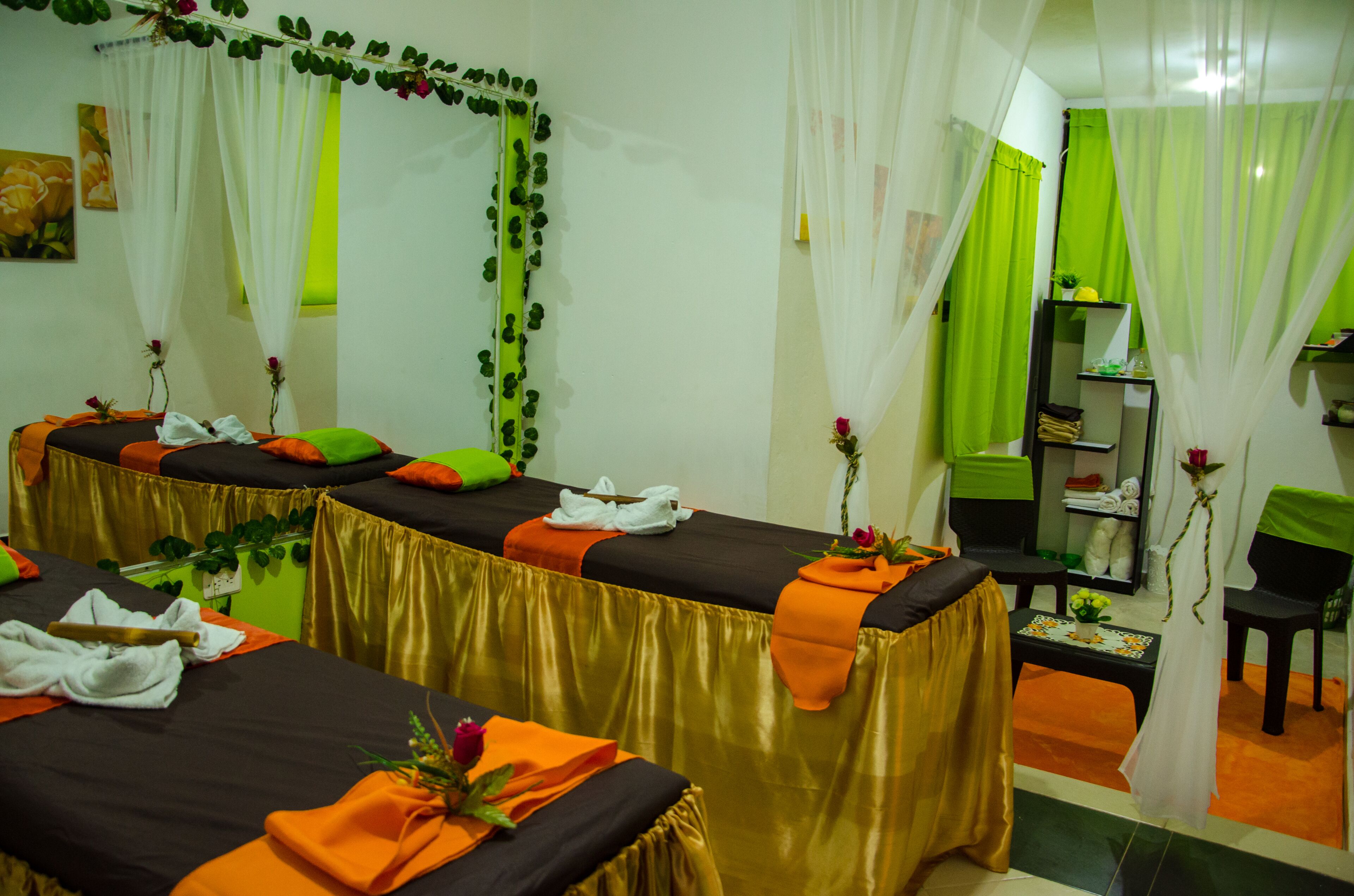 Un cuarto de spa con dos mesas de masaje, cada una con un camino dorado y toallas naranjas. Hay cortinas verdes y un espejo adornado con una guirnalda de hojas verdes.