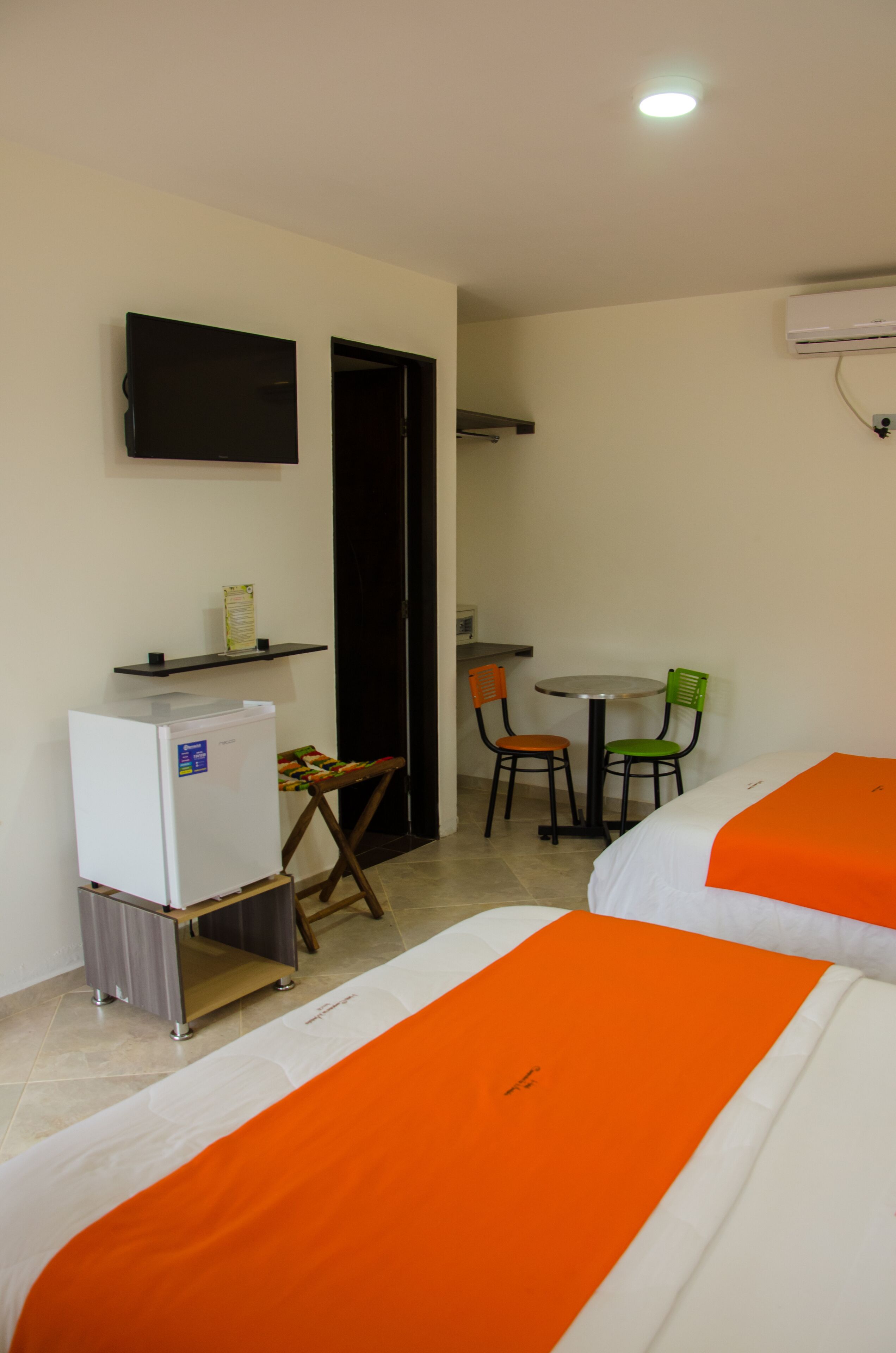 Habitación de hotel con dos camas, un escritorio pequeño con una silla, un refrigerador, un televisor montado en la pared y un equipo de aire acondicionado.