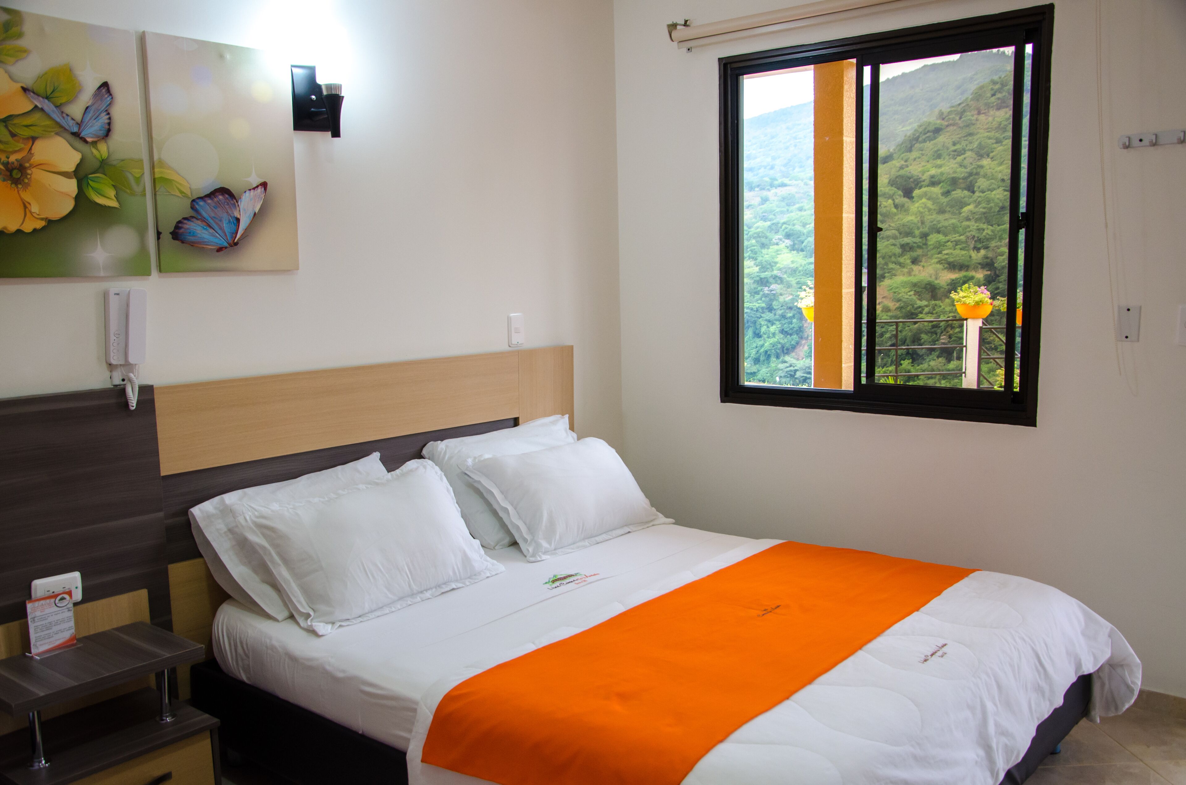 Una habitación de hotel con una cama, una mesita de noche, un teléfono en la pared, una ventana con vistas a zonas verdes y un cuadro con flores y mariposas.