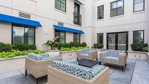 A courtyard with outdoor seating, potted plants, and a building facade with large windows and blue awnings.