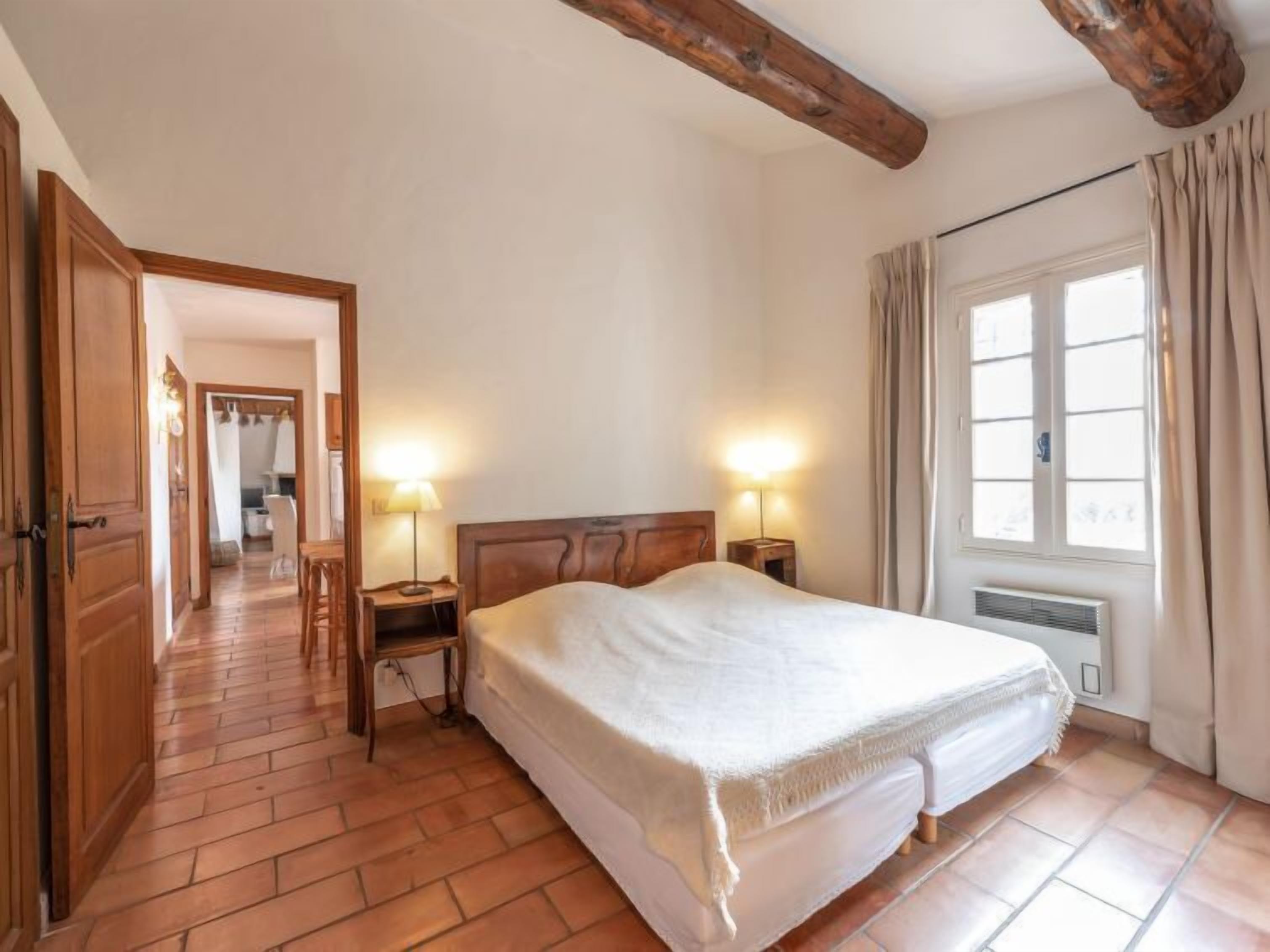 Un dormitorio con una cama grande, cabecera de madera, dos lámparas de noche, una ventana con cortinas y suelo de madera.