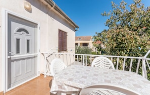 A balcony with a white table and chairs, a white door, and a view of a tree and buildings in the background.
