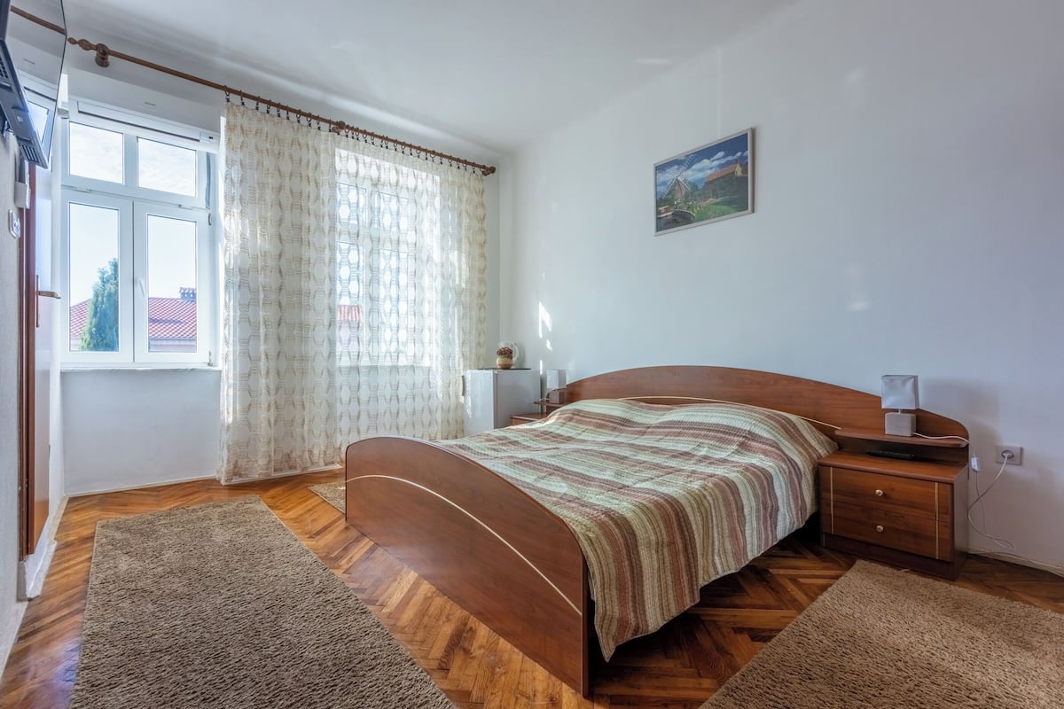 A bedroom with a wooden bed, a nightstand, a lamp, a window with curtains, and a painting on the wall.