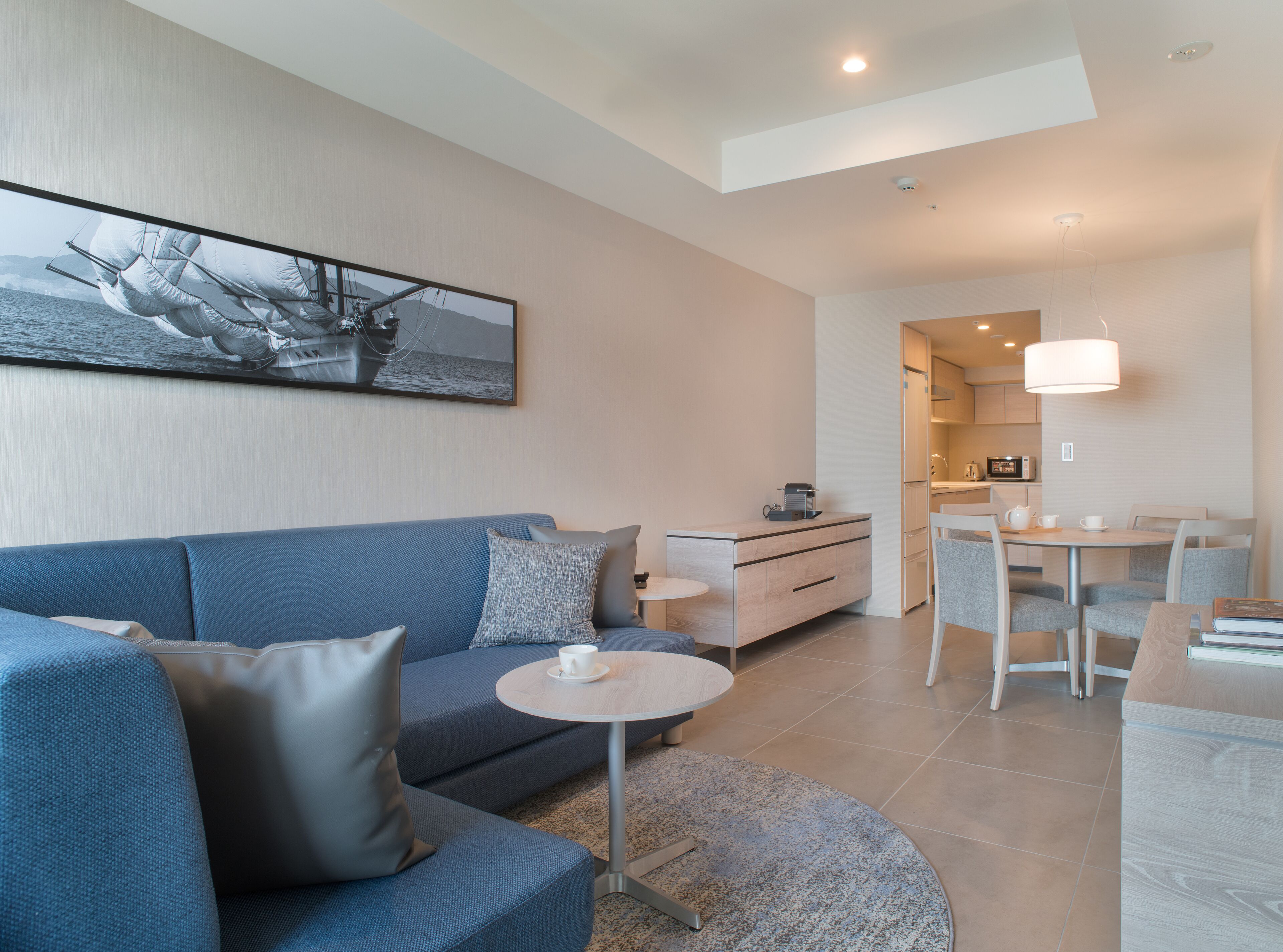 A modern living room with a blue sofa, a small round table with a cup, and a kitchen area with a dining table and chairs.
