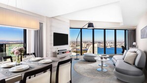A modern living room with a dining area, large windows with a city and water view, a flat-screen TV, and a sofa.