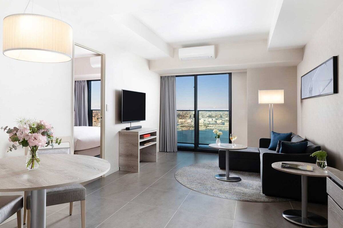 A modern hotel room with a round dining table, a sofa, a TV, and a large window with a city view.
