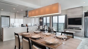 A modern dining area with a long table set for a meal, a kitchen in the background, and a large window offering a city view.