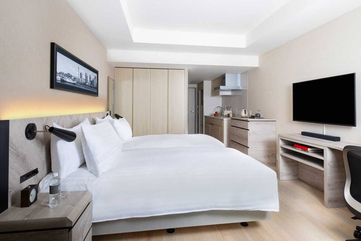 A modern hotel room with a large bed, a desk with a flat-screen TV, a chair, a small kitchenette, and a cityscape view.
